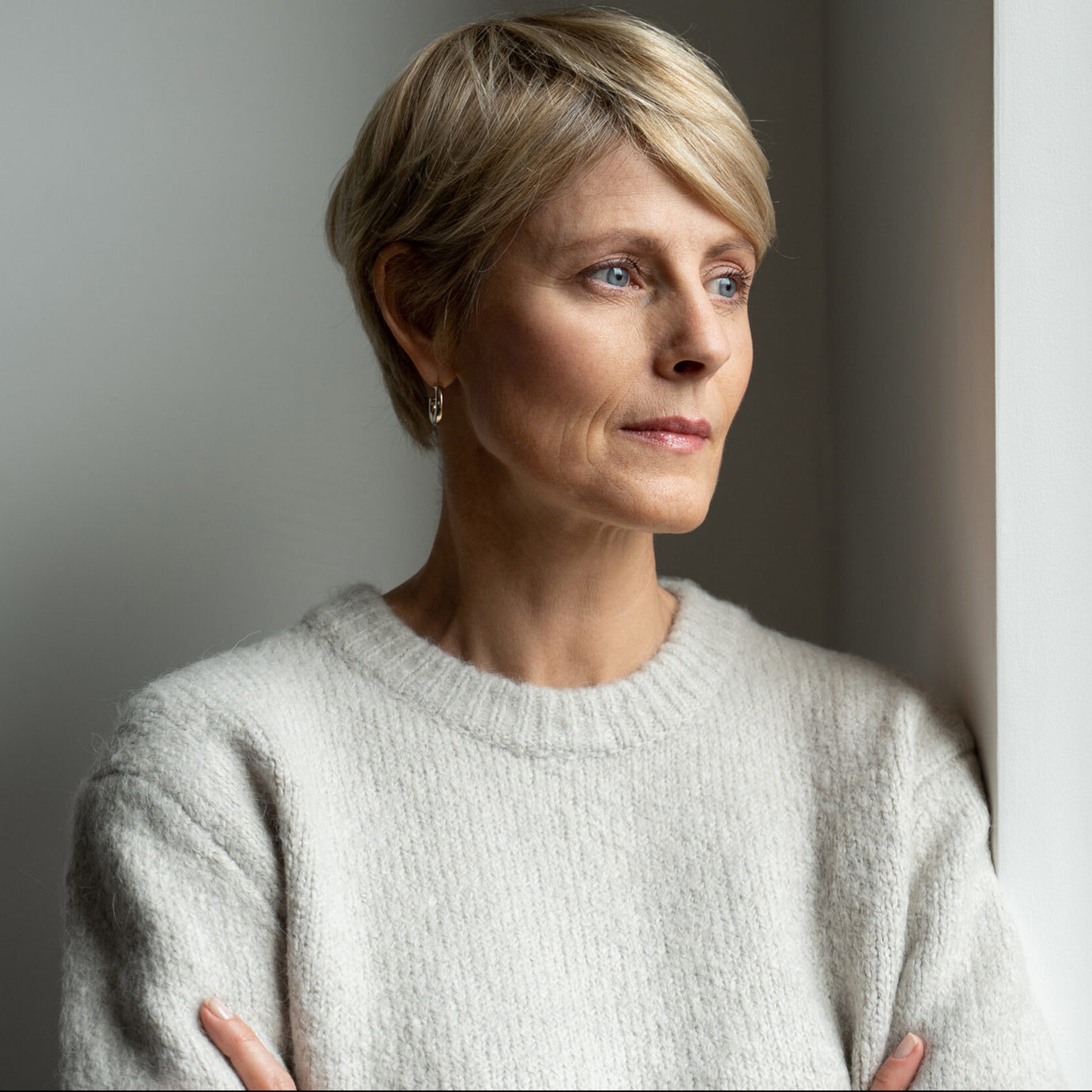 Middle-aged woman with short blonde hair wearing a grey jumper and looking thoughtfully out of a window.