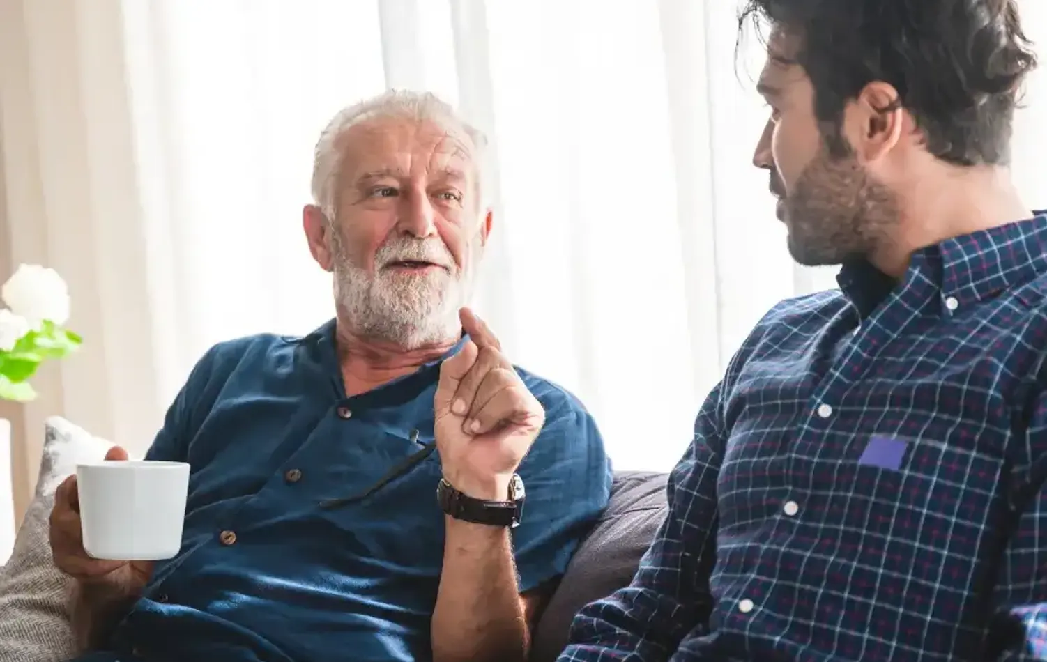 A senior man sat with a younger man on the sofa having a discussion over a coffee